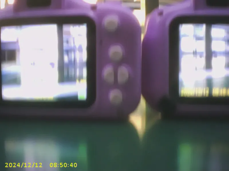 Das erste Foto auf der Karte, ein unscharfes Testbild aus der Fabrik mit zwei violetten Kinderkameras auf einem Fließband, in der Ecke der gelbe Zeitstempel 2024/12/12 08:50:40.